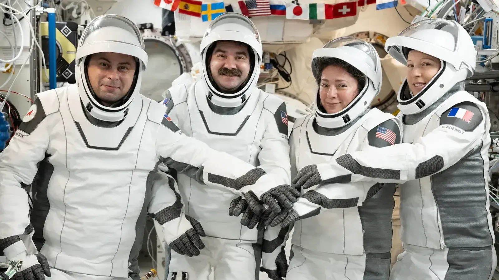 Four astronauts in white space suits smile at the camera and put their gloved hands together aboard the space station.