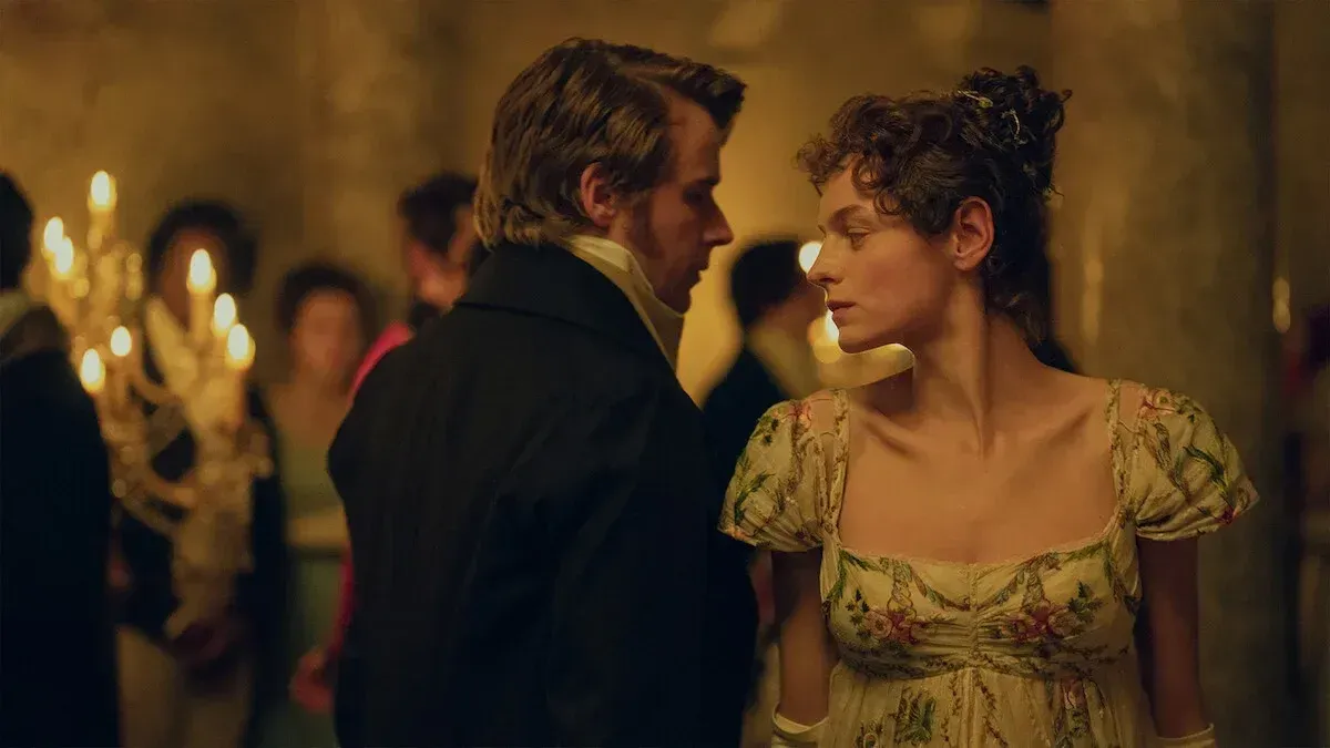 Two people dressed in historical costumes stand close together in an elegant ballroom, surrounded by softly lit chandeliers and guests, suggesting a formal or romantic setting from a past