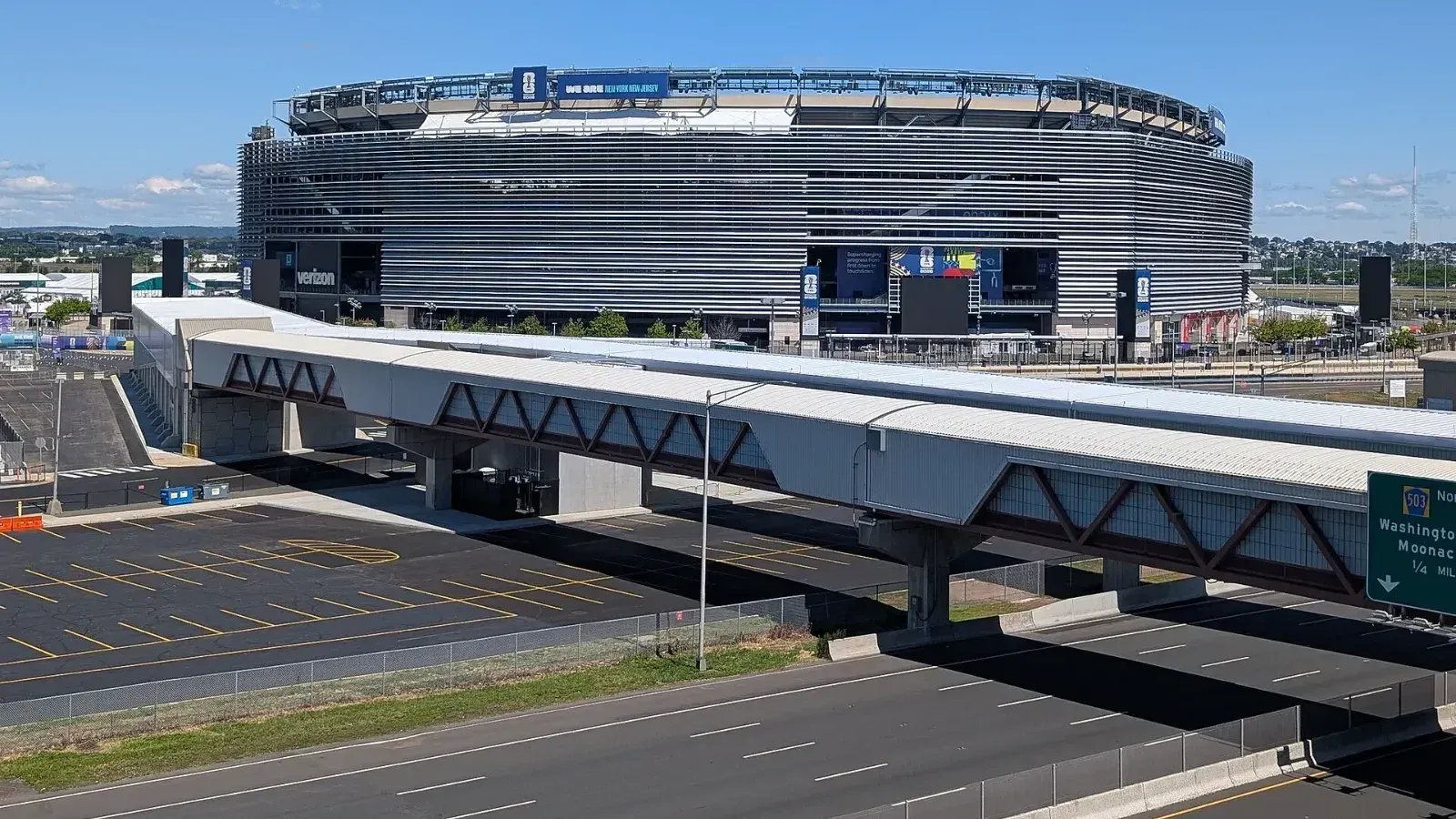 Exterior del MetLife Stadium con la decoración del Mundial 2026, visto desde un estacionamiento elevado, con una pasarela peatonal, una autopista y un estacionamiento vacío en primer plano