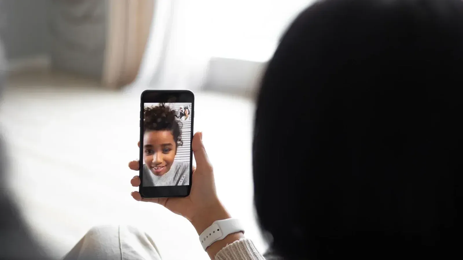 Vista posterior de una pareja anónima comunicándose con un niño afroamericano a través de video chat en un teléfono inteligente moderno mientras se sienta en la sala de estar en un fondo