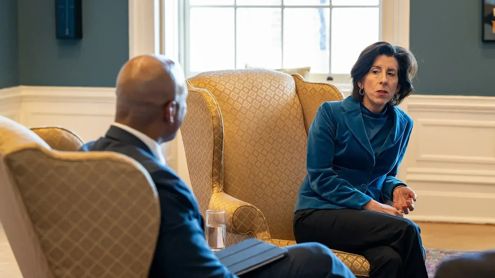 Gina Raimondo, sentada en un sillón, conversa con el gobernador de Maryland, Wes Moore, que aparece de espaldas en primer plano, en una oficina