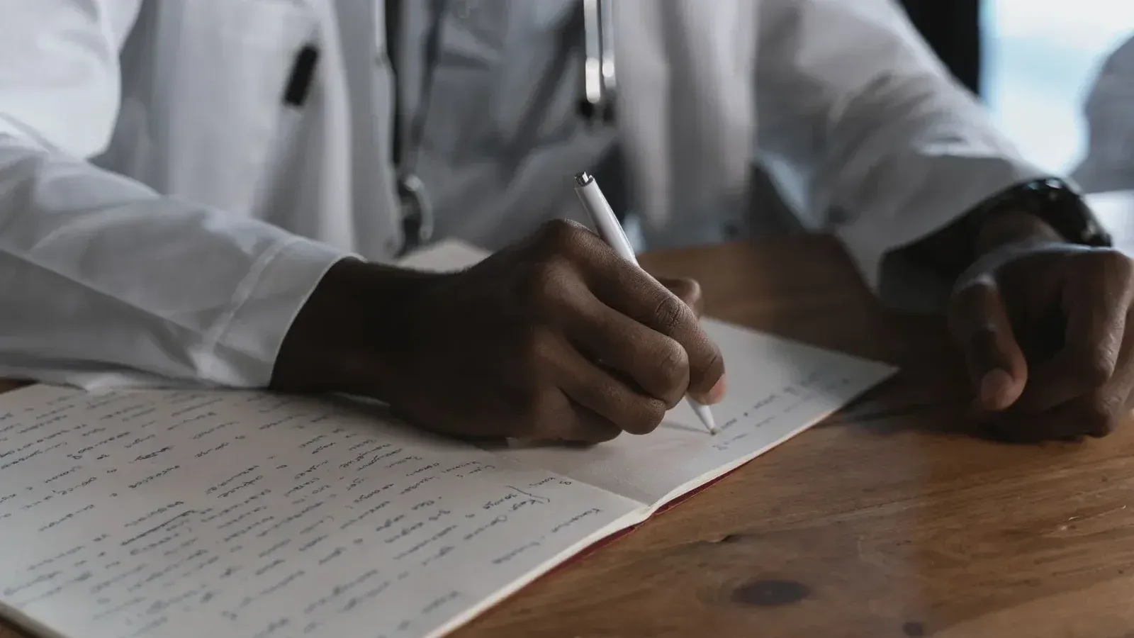 Primer plano de un médico escribiendo notas con un bolígrafo en un cuaderno médico en un escritorio de madera.