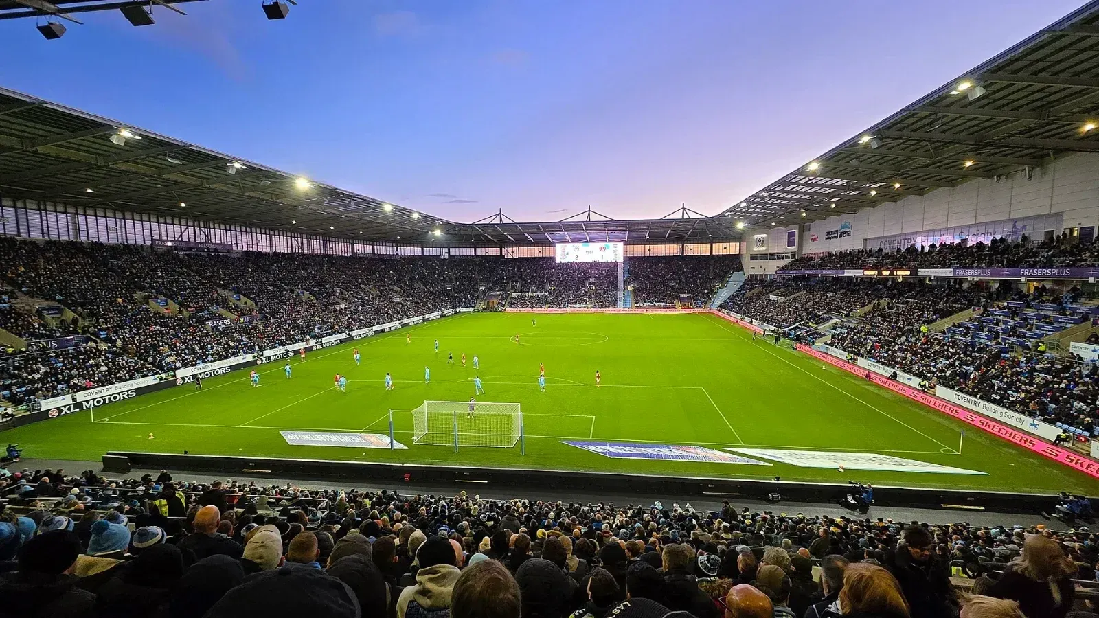 Vista desde la grada del Coventry Building Society Arena, con las tribunas llenas y los jugadores sobre el césped durante un partido al atardecer.