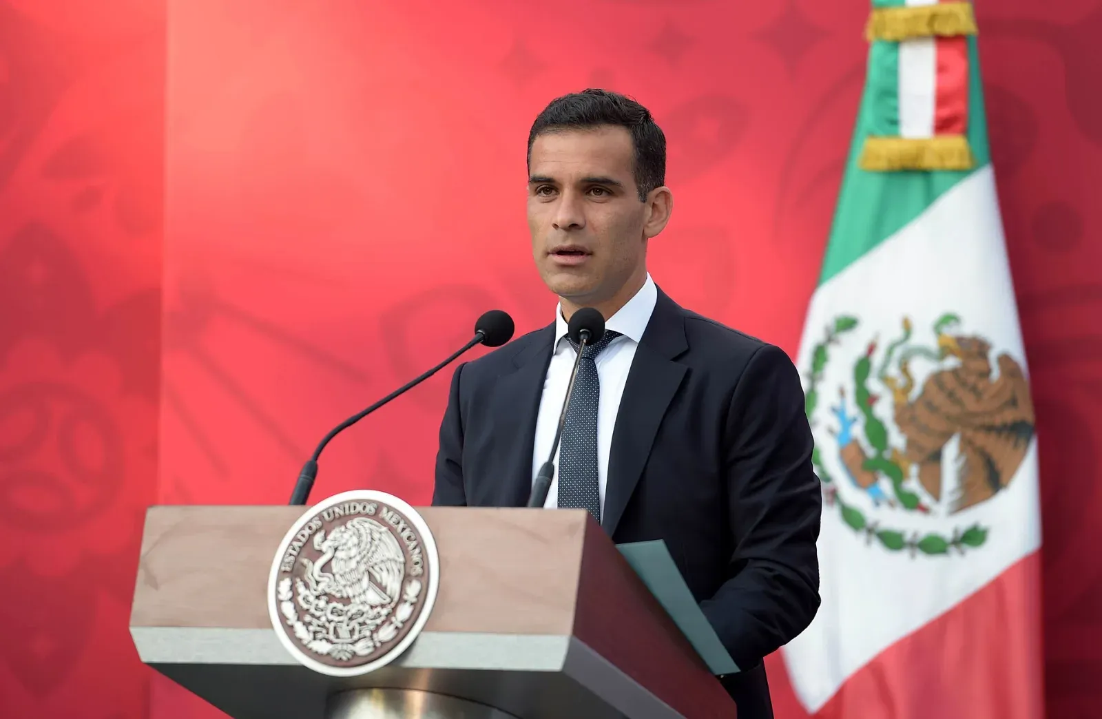 Rafael Márquez, de traje oscuro y corbata, habla ante dos micrófonos en un atril con el escudo nacional, junto a una bandera de México.