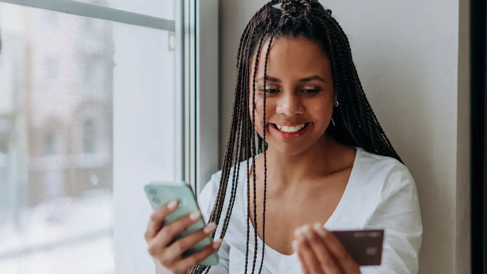 Mujer sonriente con smartphone y tarjeta de crédito para compras en línea en interiores con luz natural.