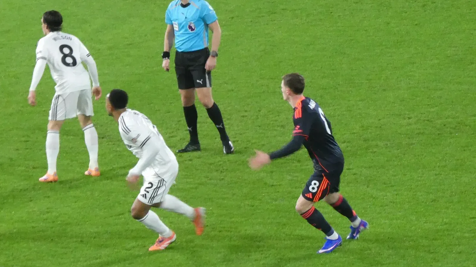 Un árbitro con camiseta celeste sigue la jugada junto a dos futbolistas de blanco y uno de azul oscuro durante un partido de la Premier League.