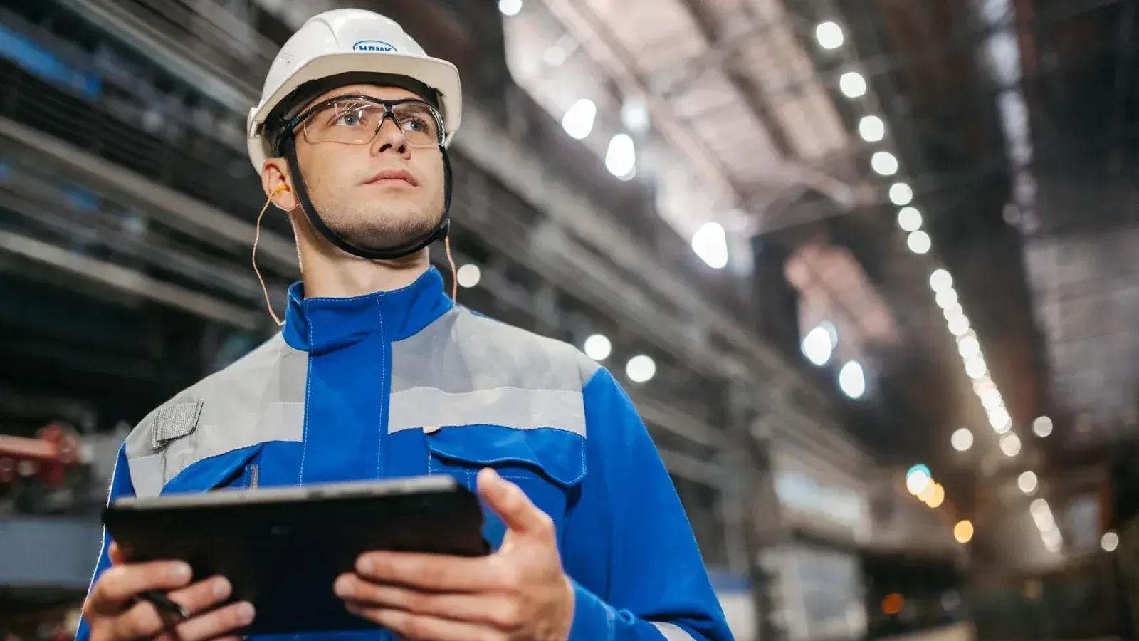 Trabajador de fábrica en equipo de seguridad usando una tableta en un entorno industrial bien iluminado.