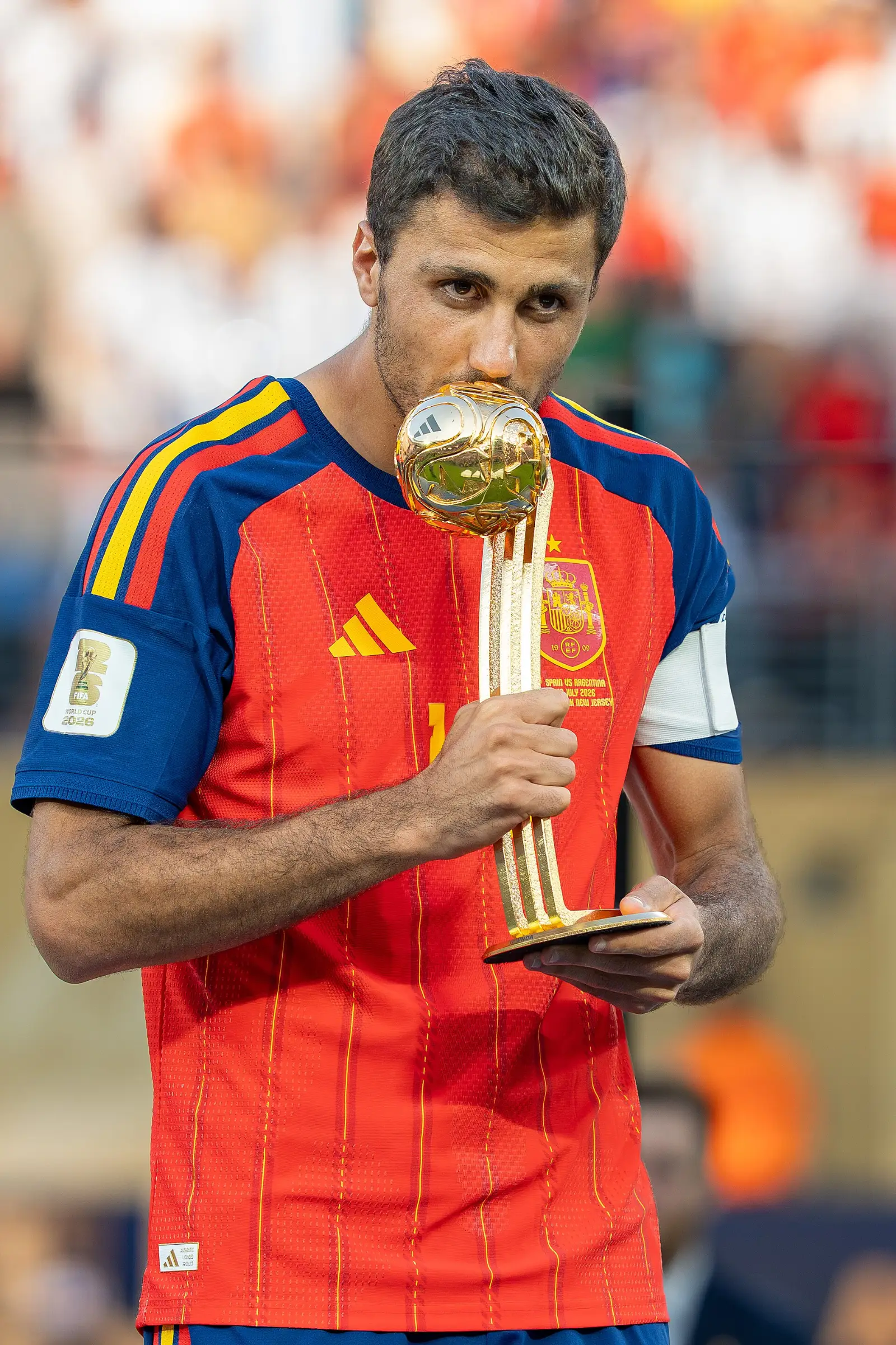 Rodri, con la camiseta roja de España y el brazalete de capitán, besa un trofeo dorado con forma de balón.