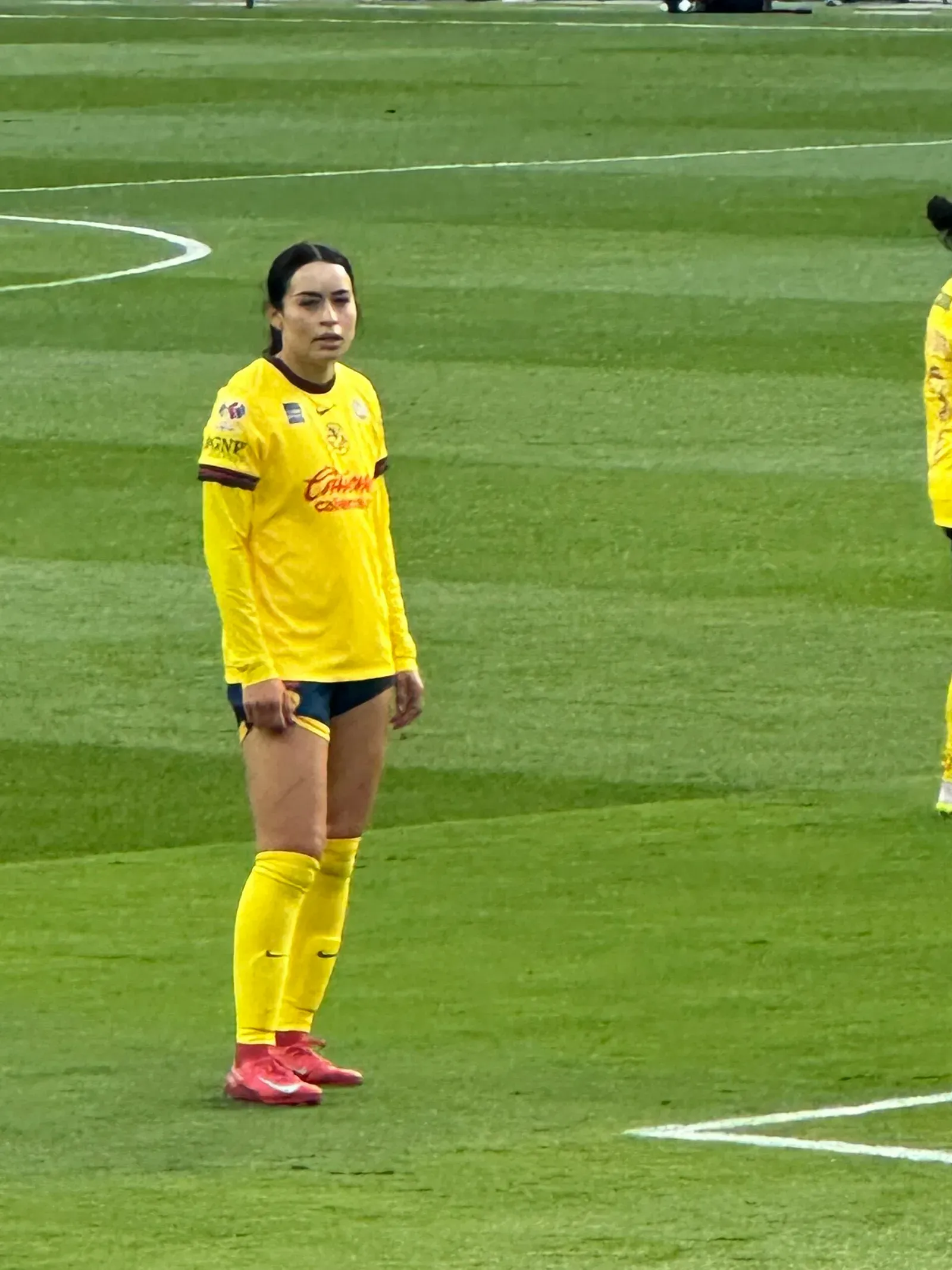 Scarlett Camberos, con la camiseta amarilla del América, short azul marino y medias amarillas, de pie sobre el césped durante un partido.