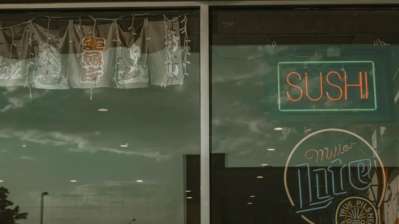 Signo de sushi de neón y reflexión en la ventana del restaurante con cielo nublado.