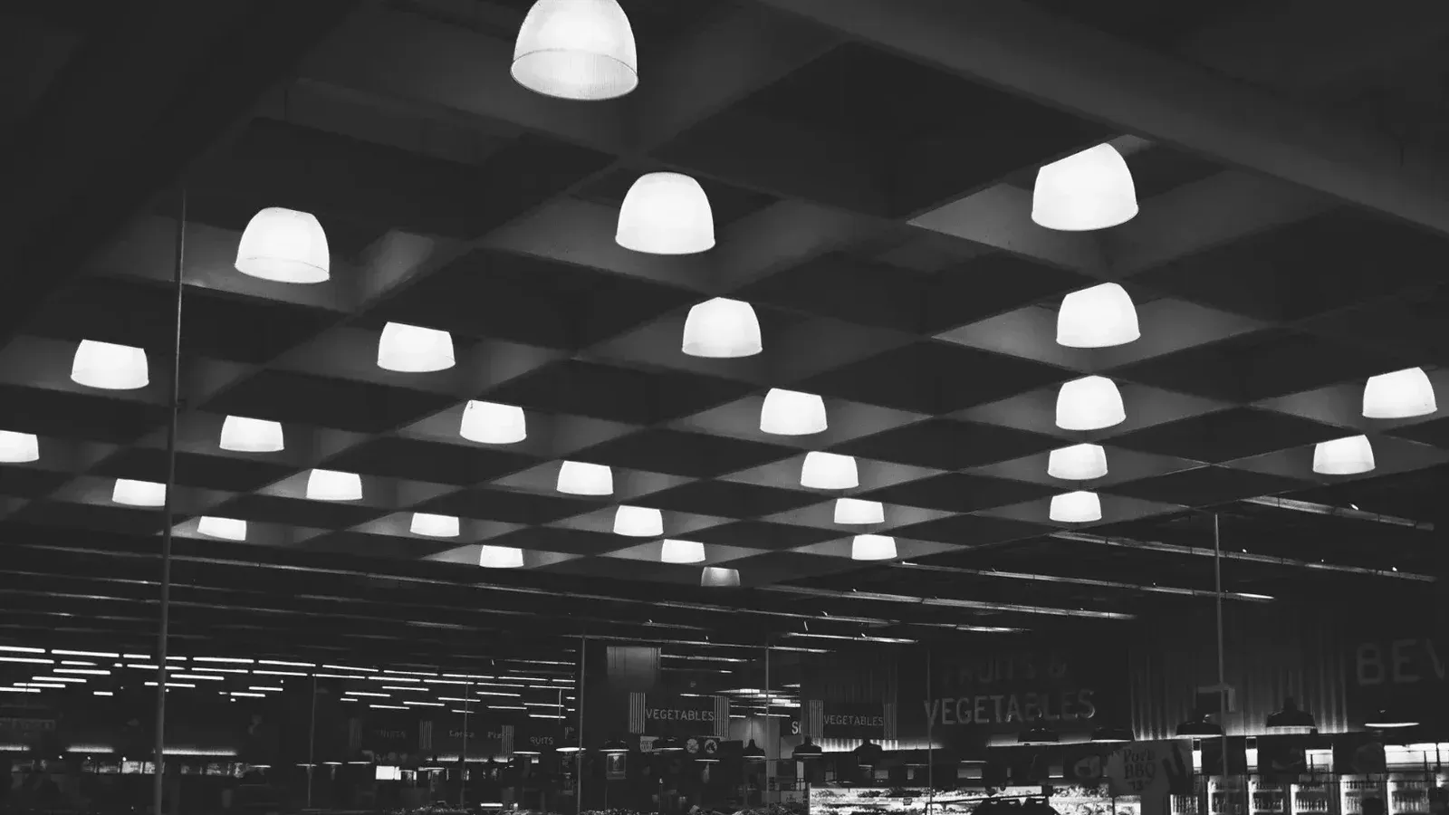Black and white of rows of glowing lamps hanging over shelves with products in modern supermarket