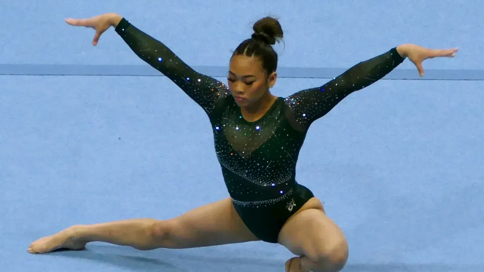 Sunisa Lee, con maillot verde oscuro de manga larga, se agacha con una pierna flexionada, la otra estirada y los brazos extendidos sobre el tapiz azul en su ejercicio de suelo