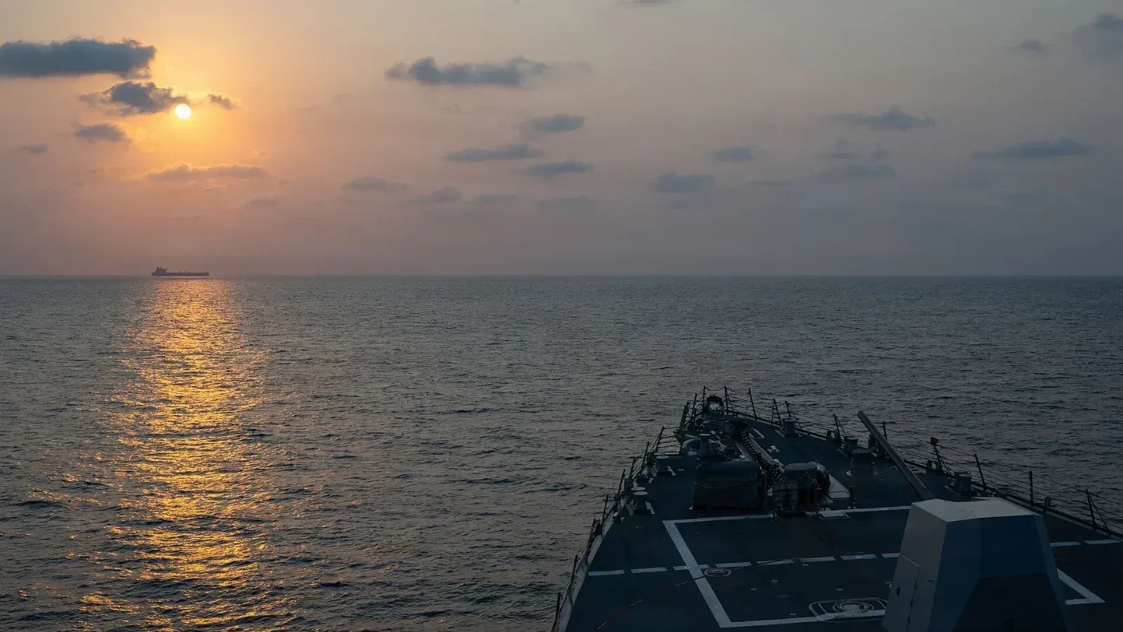 Cubierta de proa de un destructor de la Marina de Estados Unidos, con su cañón, en mar abierto; a lo lejos, un buque mercante se recorta en el horizonte con el sol bajo.