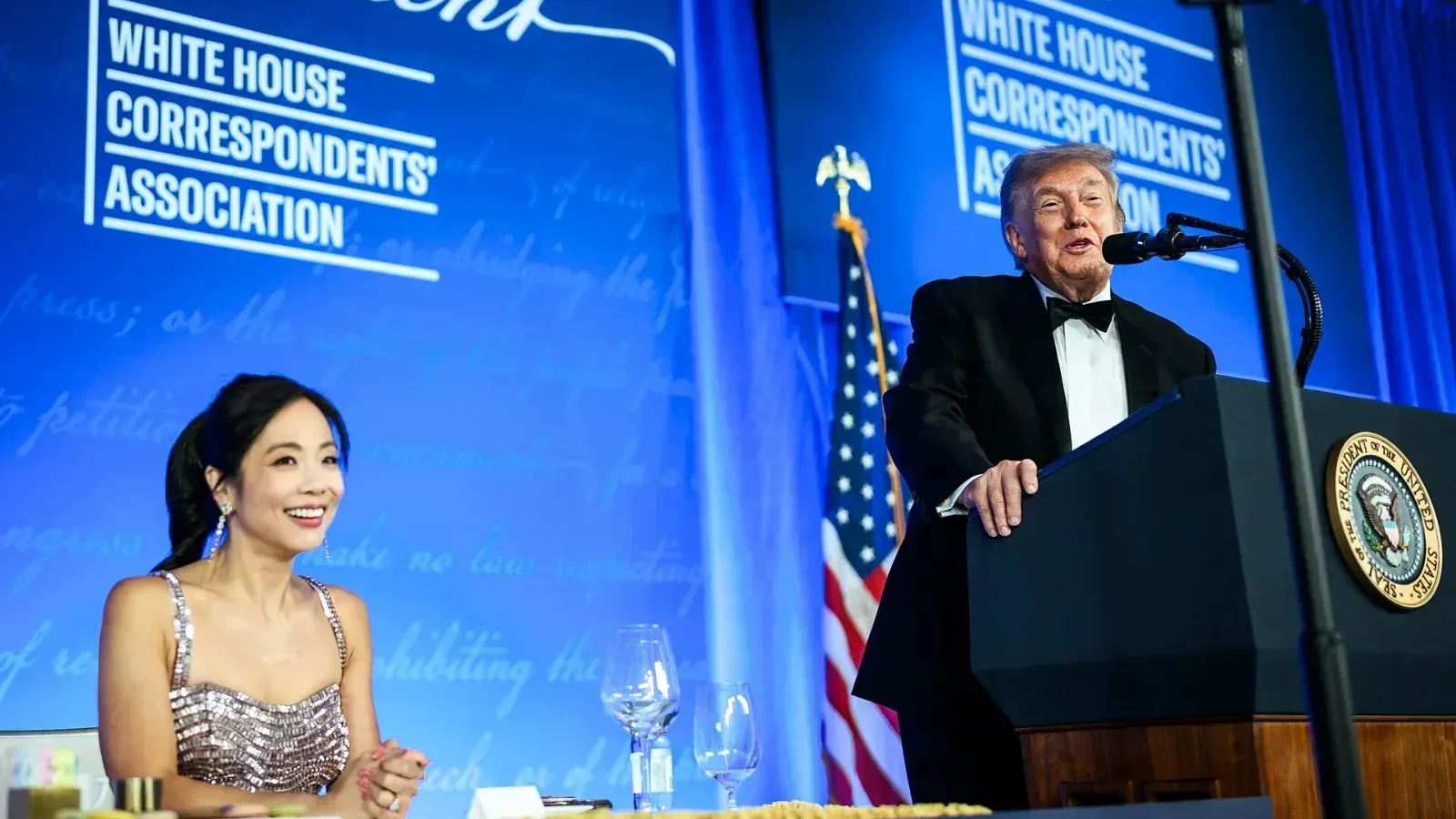 Donald Trump, de esmoquin y pajarita, habla en un atril con el sello presidencial; a su lado, una mujer con vestido de lentejuelas sonríe sentada a la mesa.