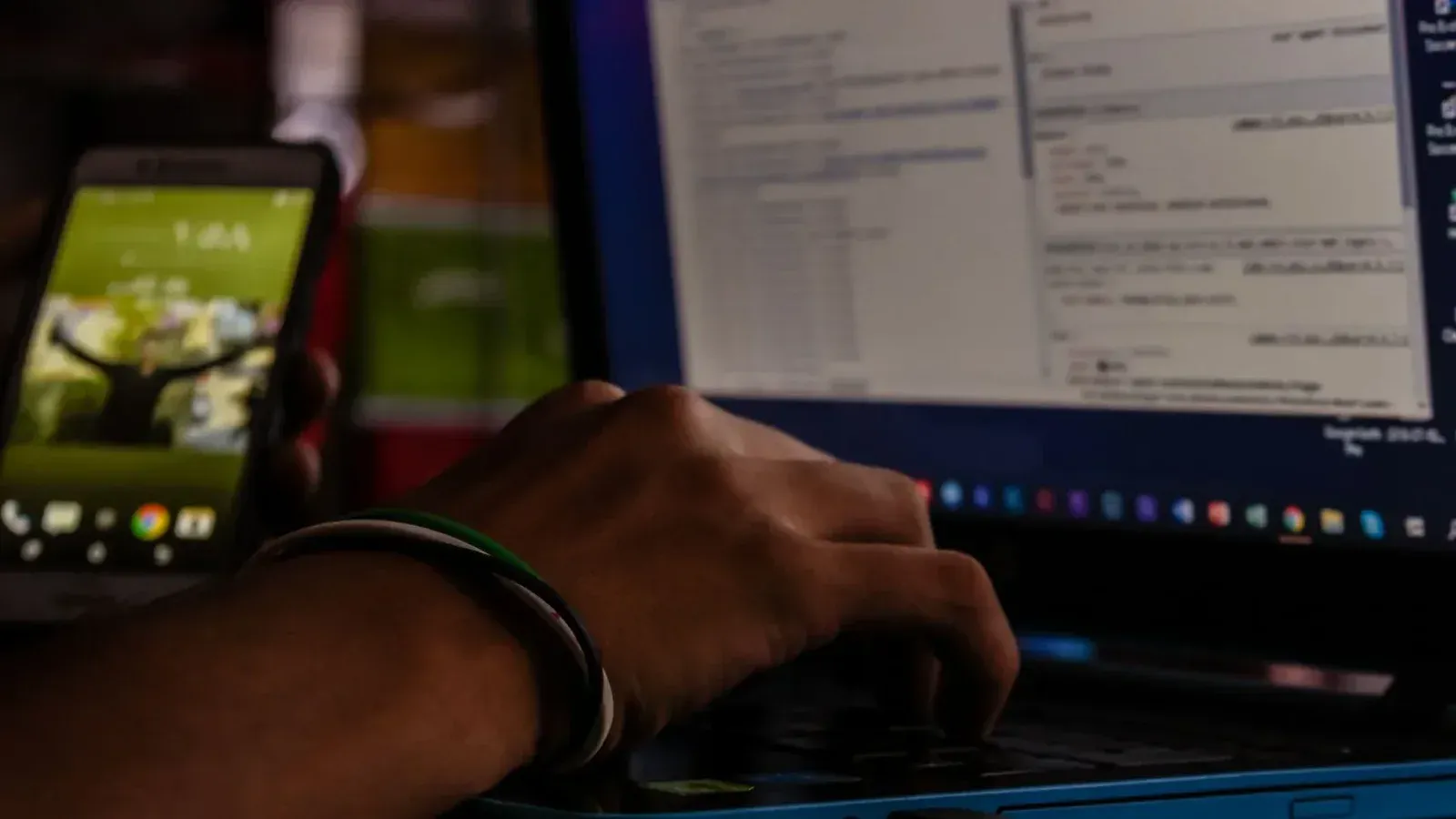 Close-up of a person working on a laptop and smartphone in a dimly lit office, showcasing remote work.