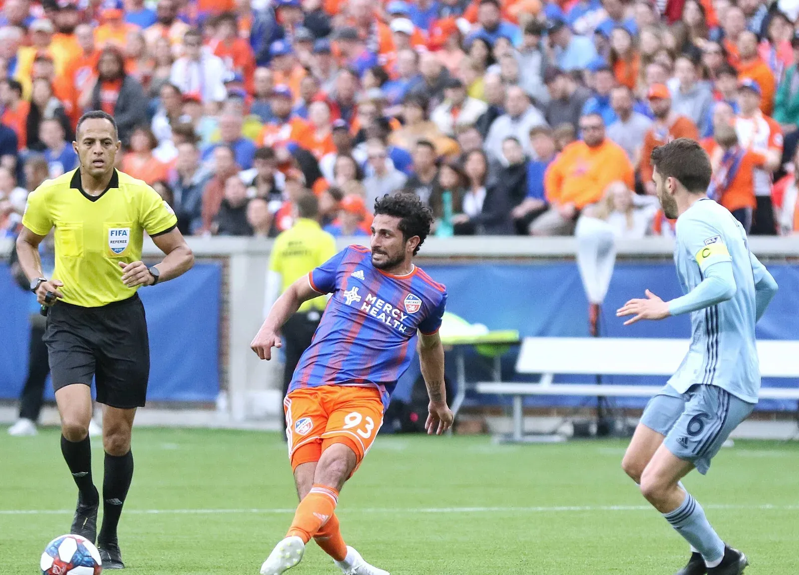 Un árbitro de amarillo con la insignia de la FIFA sigue la jugada mientras un futbolista de camiseta azul y naranja pasa el balón ante un rival de celeste.