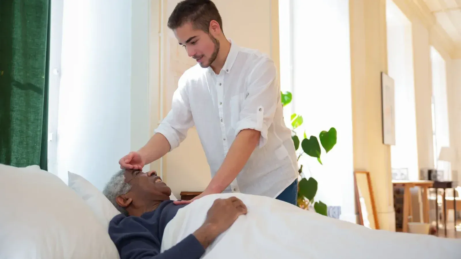 Un joven apoya a un anciano en la cama, ofreciendo cuidado en una habitación luminosa.