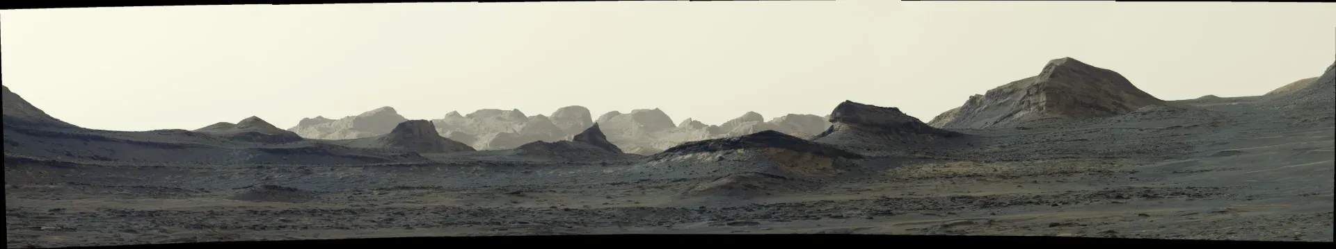 Una amplia vista panorámica de un paisaje marciano grisáceo y de color canela grisáceo presenta formaciones rocosas irregulares y crestas montañosas onduladas bajo un cielo brumoso. Las
