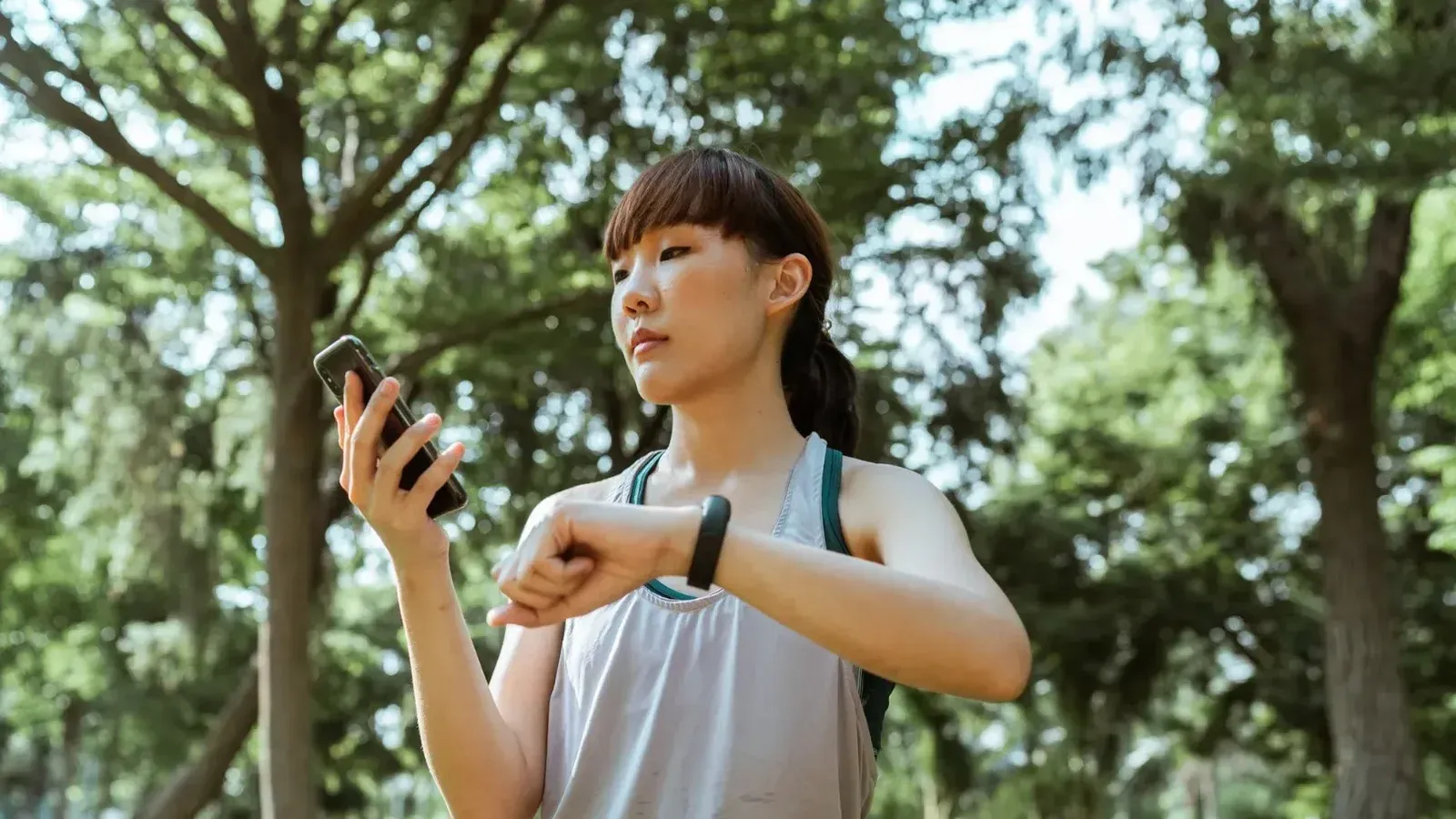 Joven deportista asiática en forma positiva en forma activa en smartphone de navegación activa y comprobación de pulsera de fitness mientras entrena en parque soleado verde