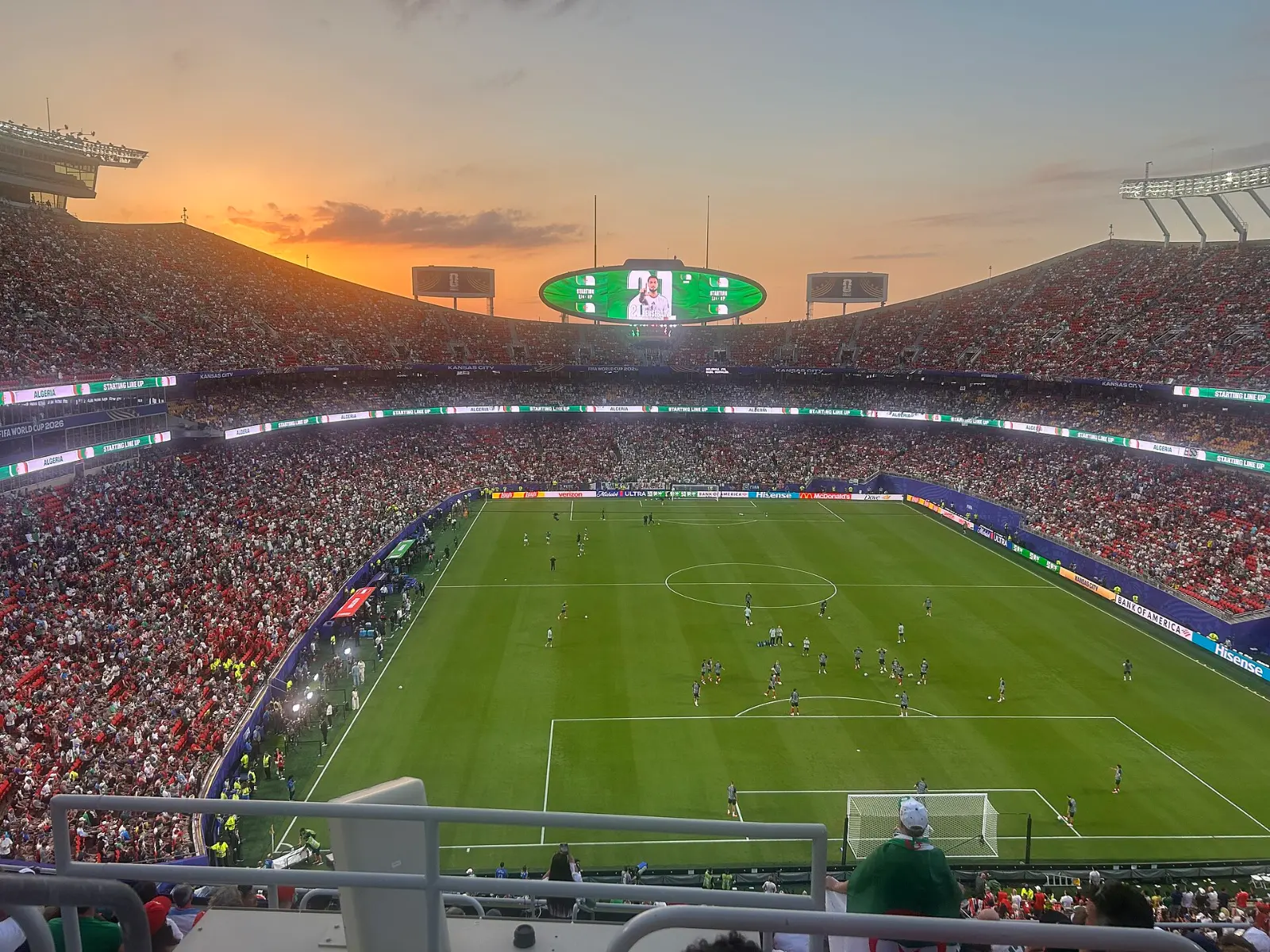 Vista desde la grada alta de un estadio lleno al atardecer, con jugadores sobre el césped y una pantalla ovalada suspendida sobre el campo.