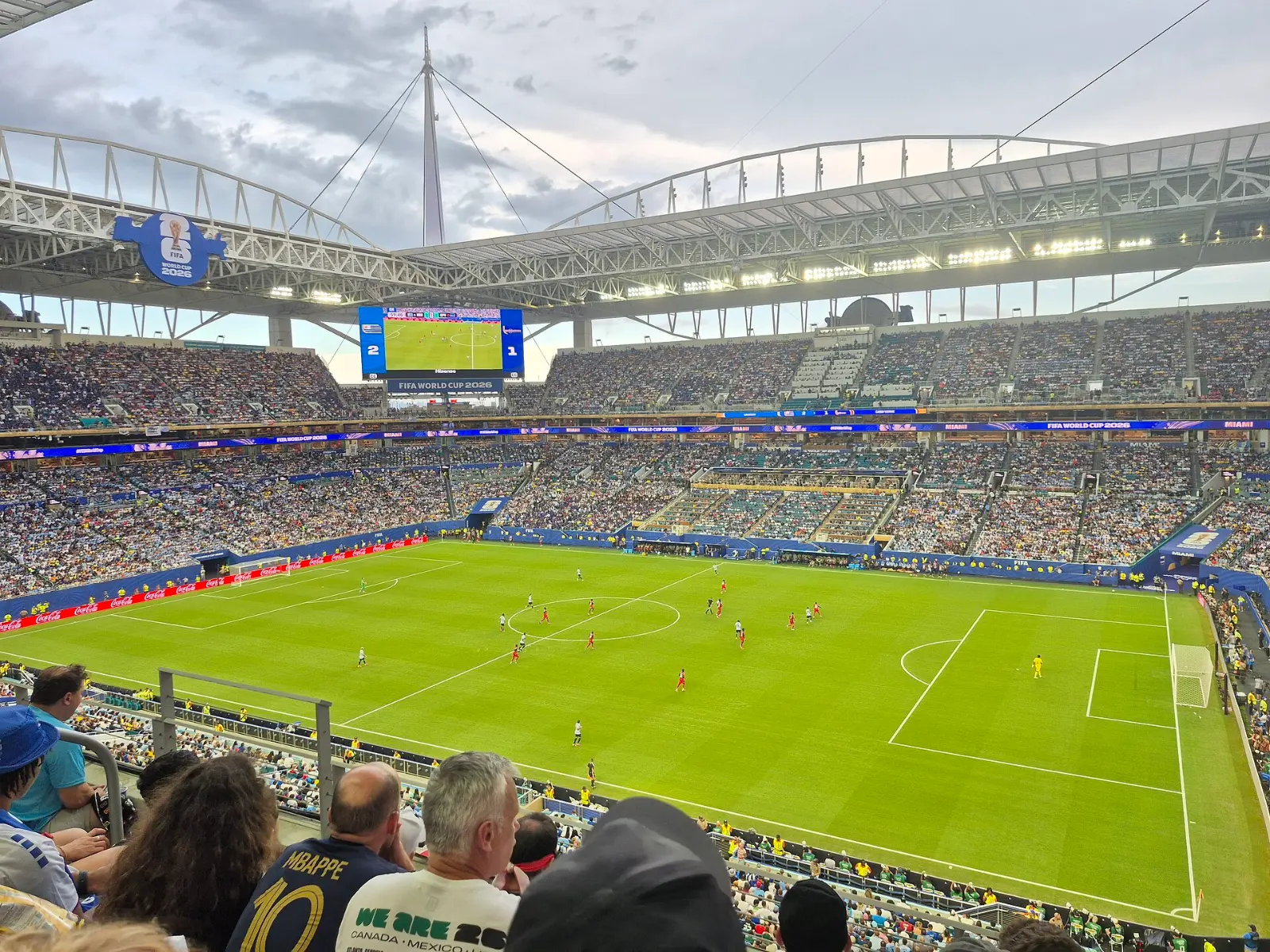Vista desde la tribuna de un estadio lleno del Mundial 2026, con jugadores de rojo y de blanco en la cancha y el marcador 2-1 en la pantalla gigante.