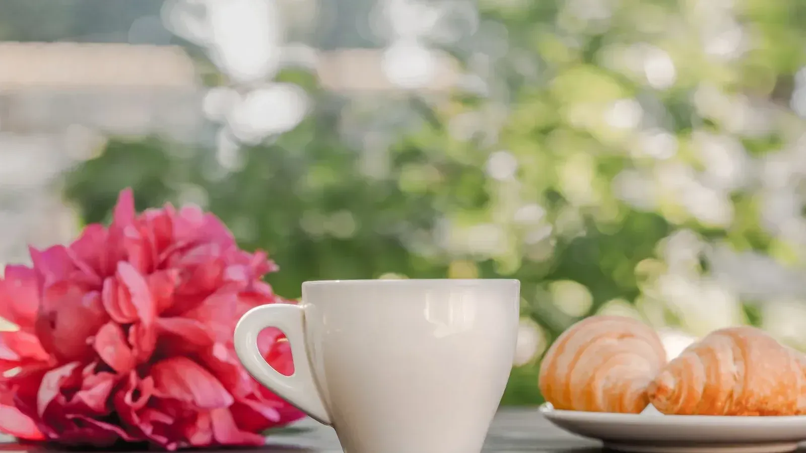 Taza de café caliente servido en mesa de madera con manojo de peonías rosas y plato de croissants recién horneados durante el desayuno en la terraza en la mañana soleada