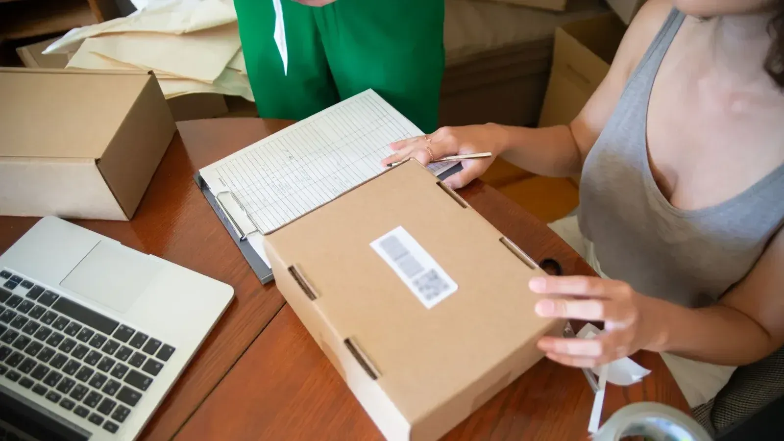 Dos personas empacando pedidos en línea en un entorno de pequeña empresa con una computadora portátil.