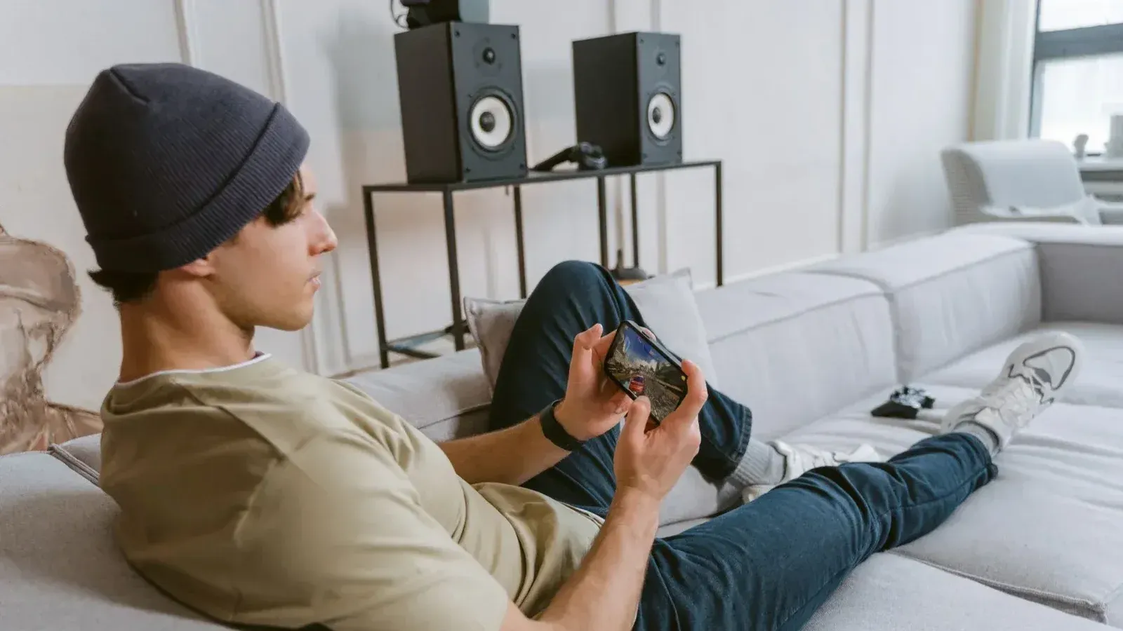 Joven casual jugando juegos móviles en el sofá con altavoces en el fondo.