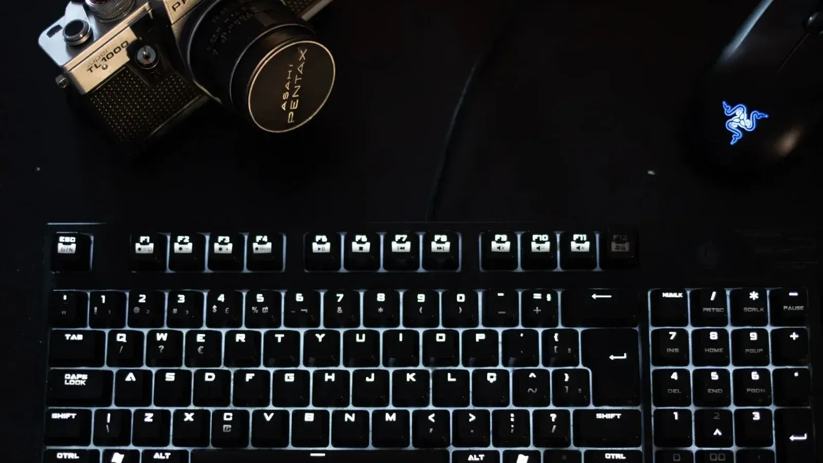 Una configuración tecnológica con poca luz que incluye un teclado iluminado, una cámara vintage y un mouse moderno.