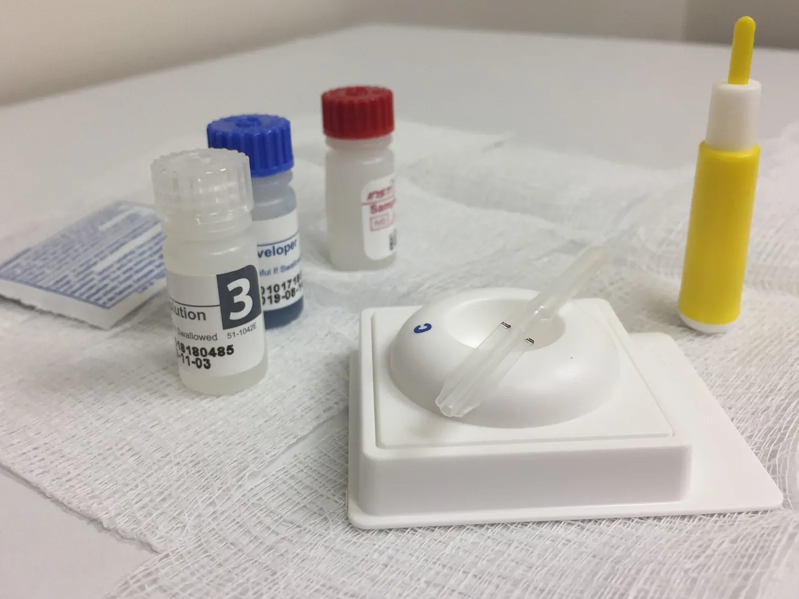 Kit de prueba rápida sobre una gasa: tres frascos pequeños con tapas blanca, azul y roja, un dispositivo de plástico blanco con una pipeta y una lanceta amarilla.