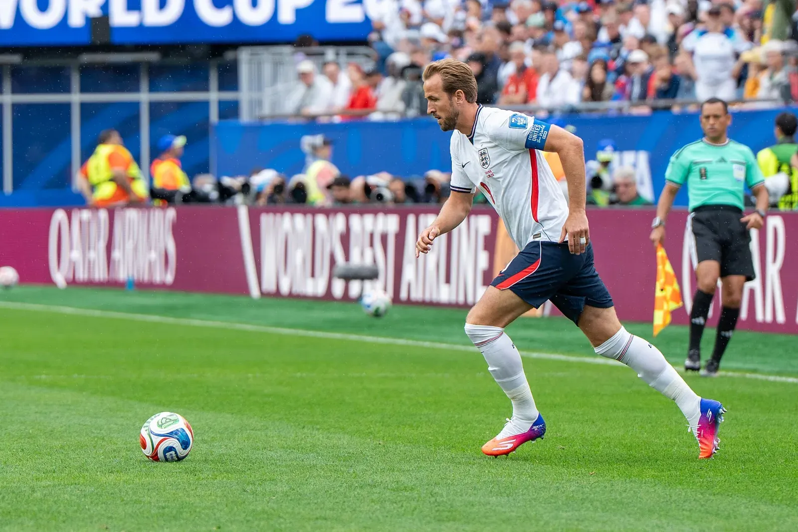 Harry Kane, con la camiseta blanca de Inglaterra y el brazalete de capitán, corre hacia el balón sobre el césped; detrás, un asistente arbitral con su bandera.