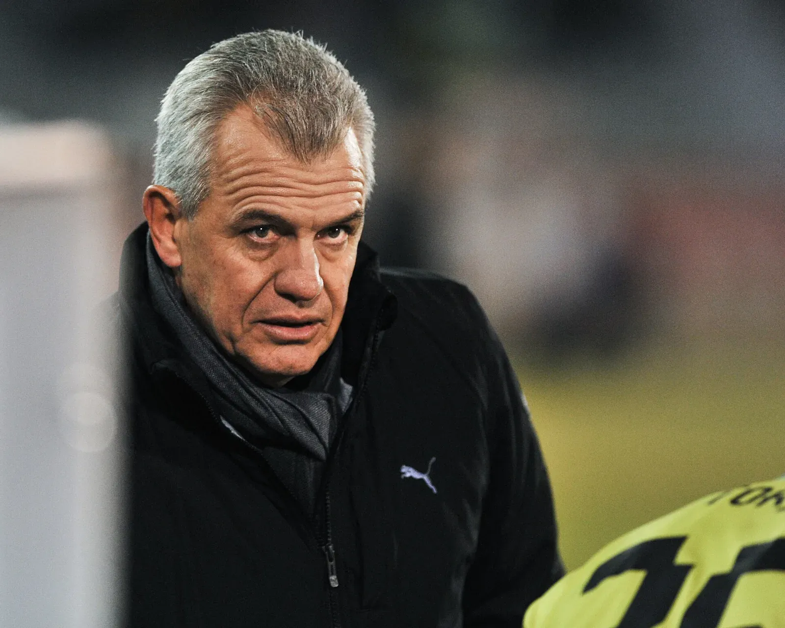 Javier Aguirre, de cabello canoso, con chaqueta negra y bufanda gris, mira al frente con gesto serio; a la derecha asoma el hombro de un jugador con camiseta amarilla.