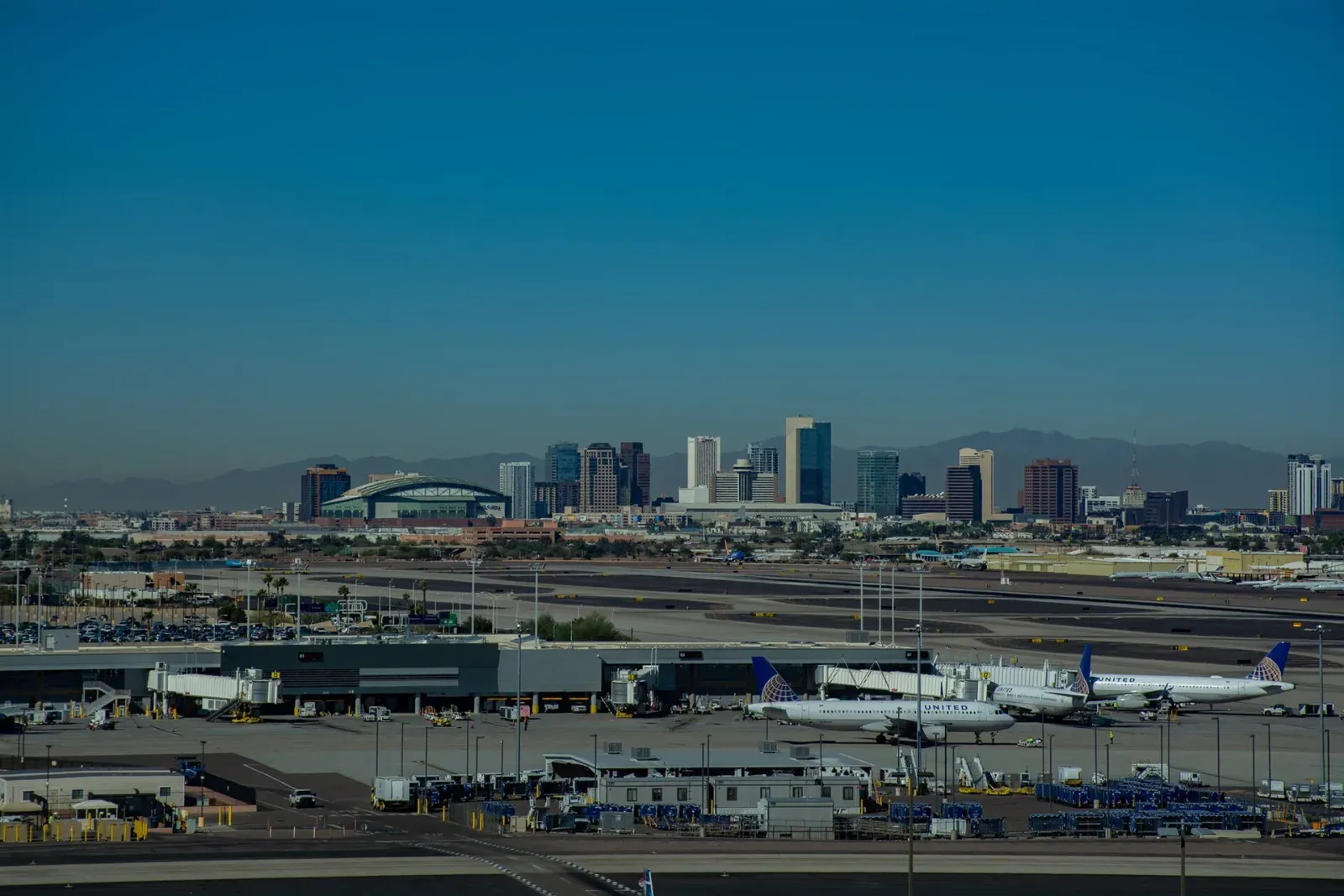 Rascacielos del centro de Phoenix al fondo, con montañas detrás, y en primer plano la pista y una terminal del aeropuerto con aviones de United estacionados.