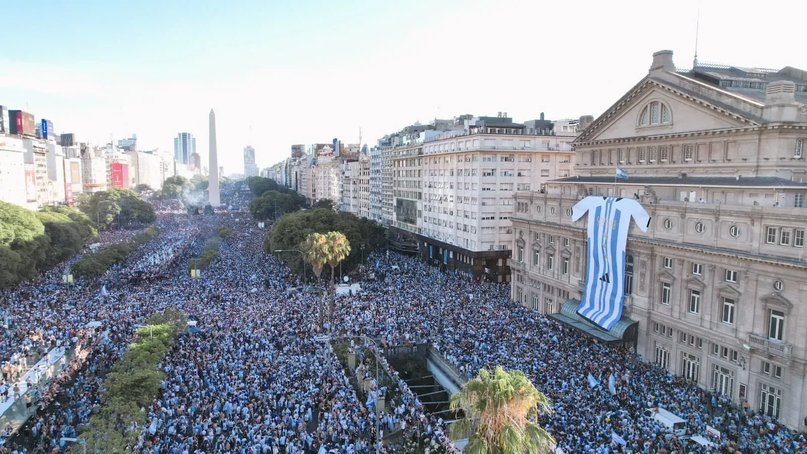 Vista aérea de una multitud vestida de celeste y blanco que llena una amplia avenida, con el Obelisco al fondo y una camiseta gigante de Argentina colgada en la fachada de un edificio.