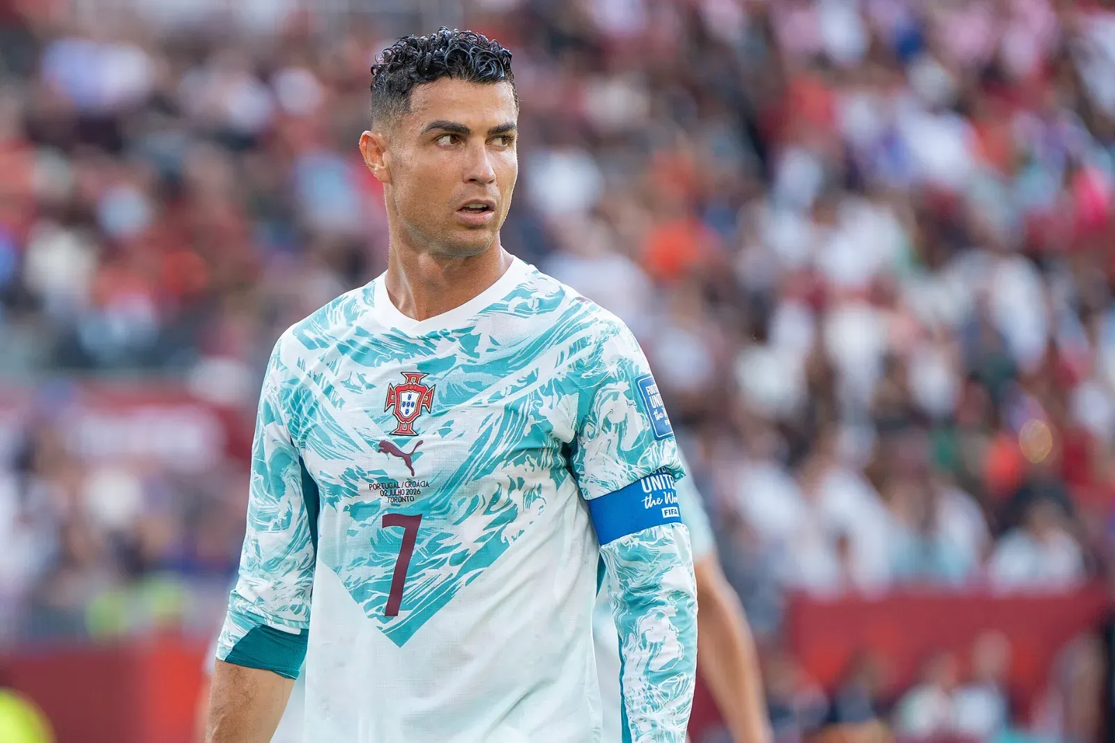 Cristiano Ronaldo, con la camiseta blanca y turquesa de Portugal, el dorsal 7 y el brazalete de capitán, mira hacia un lado con gesto serio ante la grada.