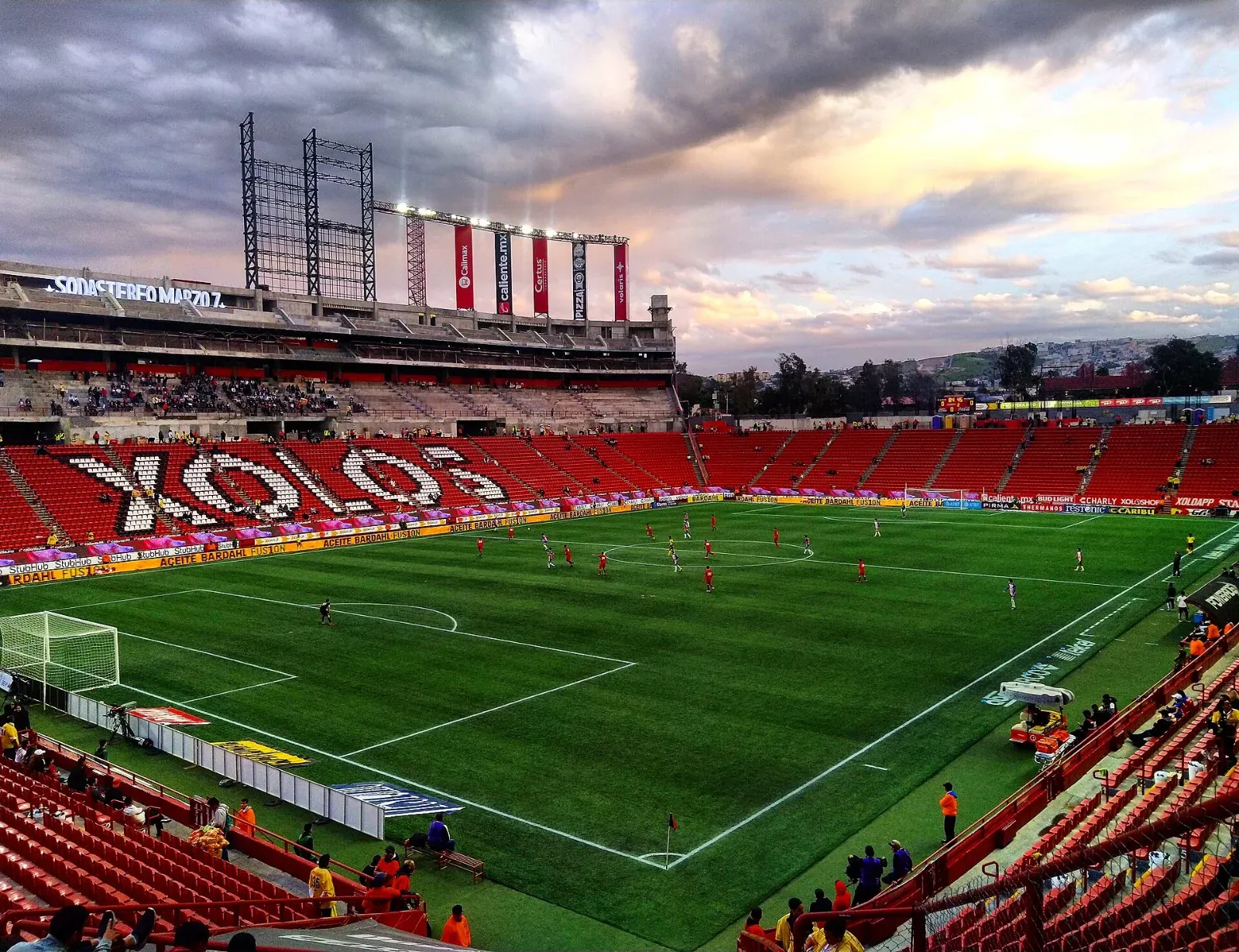 Vista del Estadio Caliente de Tijuana durante un partido, con la palabra XOLOS formada en las gradas rojas, jugadores en la cancha y un cielo nublado al atardecer.