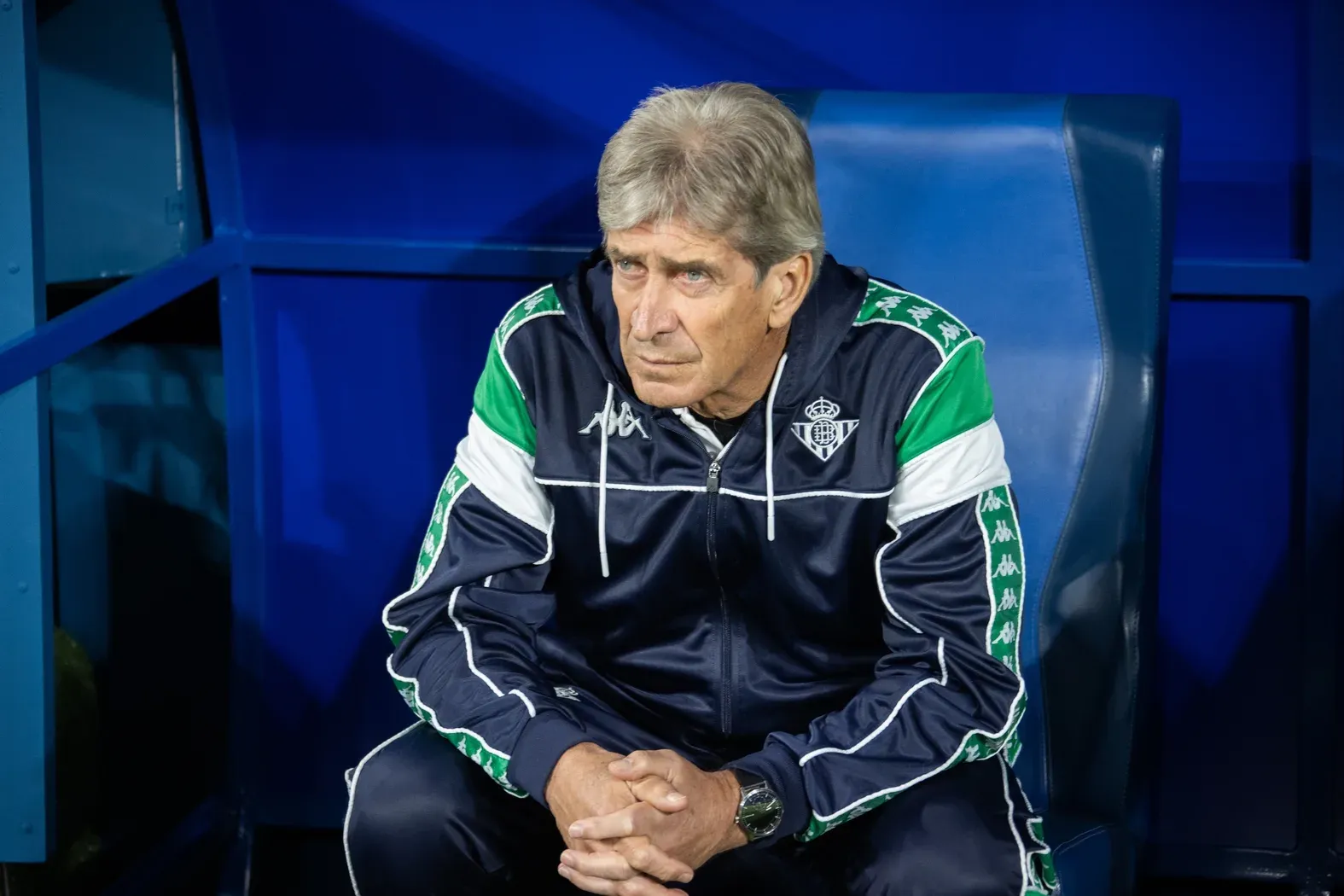 Manuel Pellegrini, de cabello canoso y chaqueta azul marino del Betis con franjas verdes y blancas, sentado en el banquillo con las manos entrelazadas.