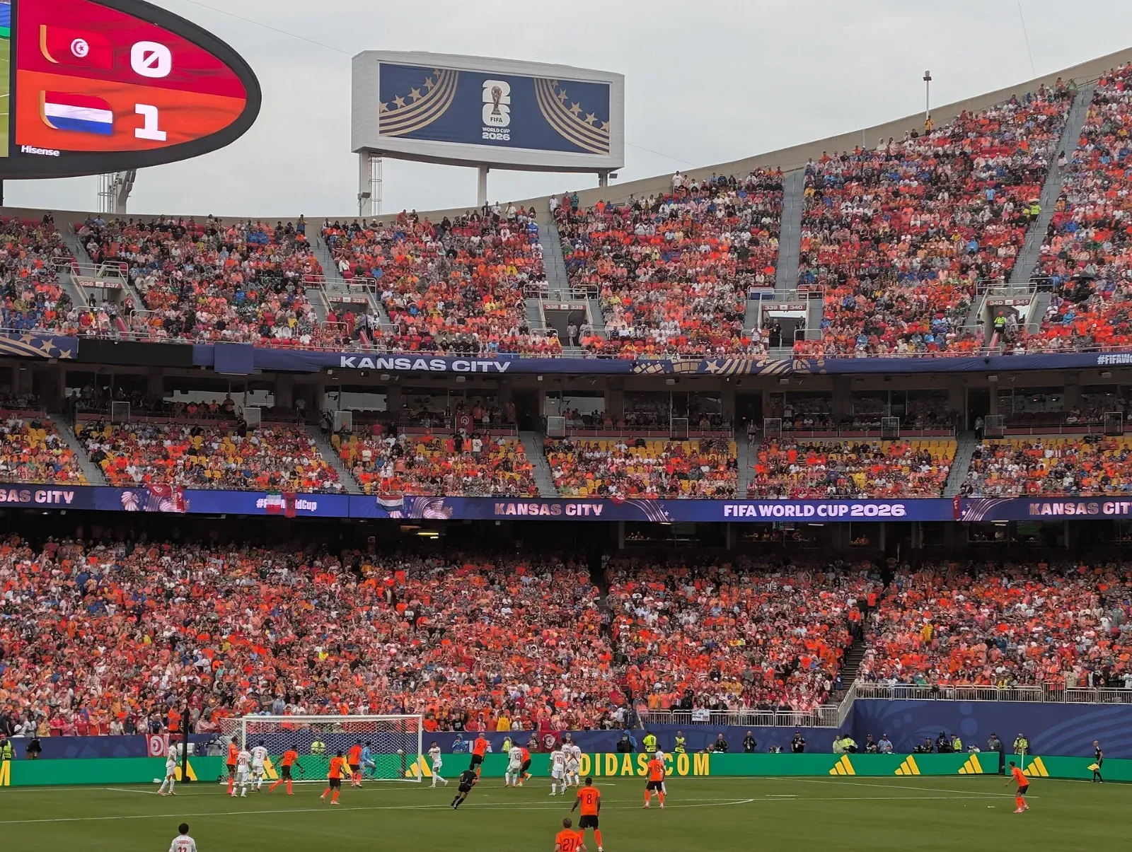 Estadio lleno de aficionados vestidos de naranja; el marcador indica Túnez 0 y Países Bajos 1 mientras los jugadores disputan una jugada cerca de la portería.