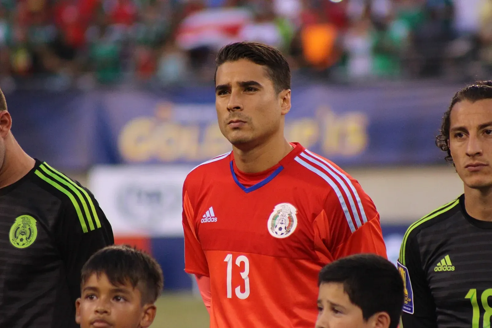 Guillermo Ochoa, con la camiseta roja de portero de México y el número 13, posa serio en el campo junto a compañeros de uniforme negro y niños acompañantes.