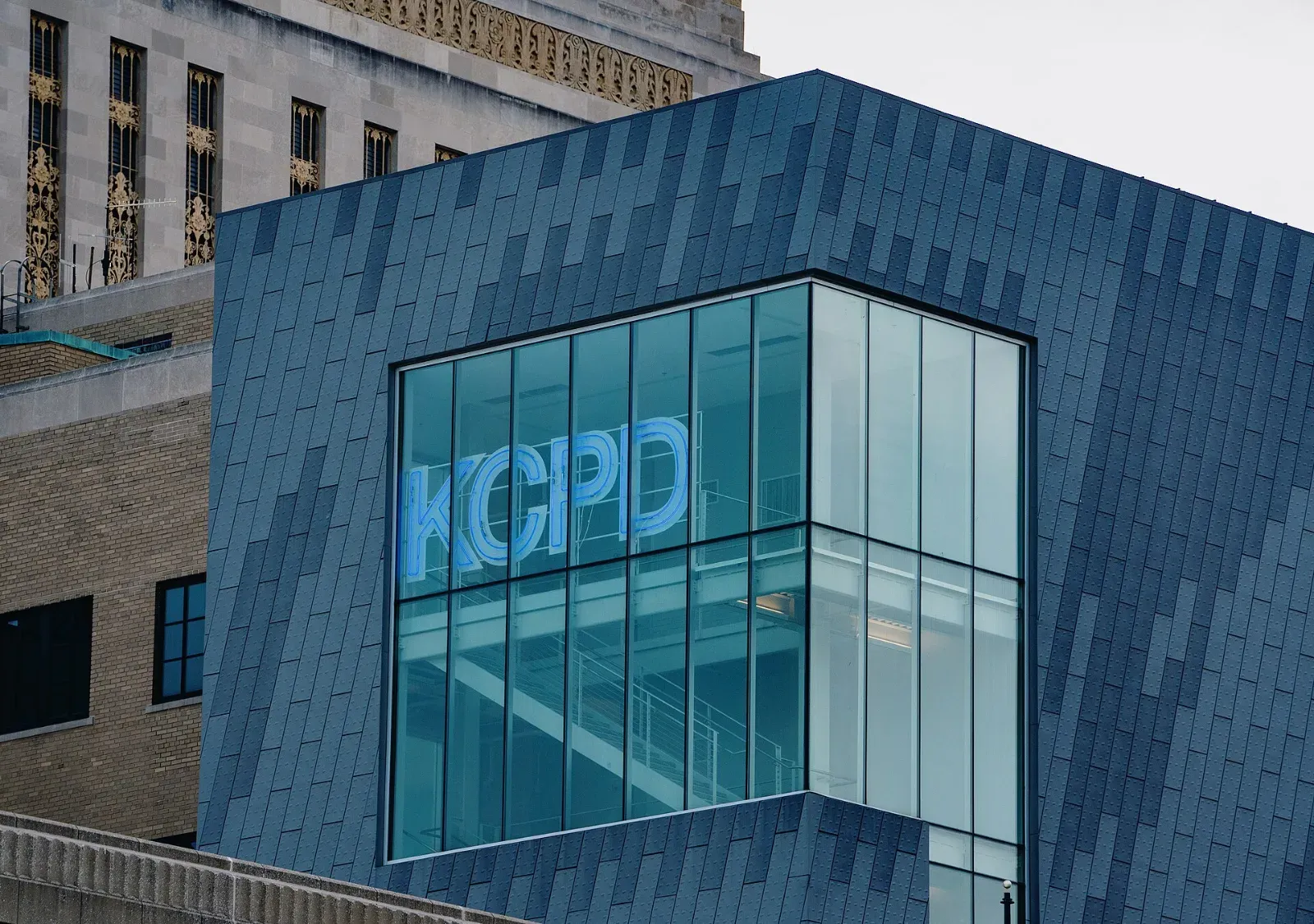Fachada de paneles grises y vidrio de la sede de la policía de Kansas City, con el letrero luminoso azul «KCPD» detrás de un ventanal.