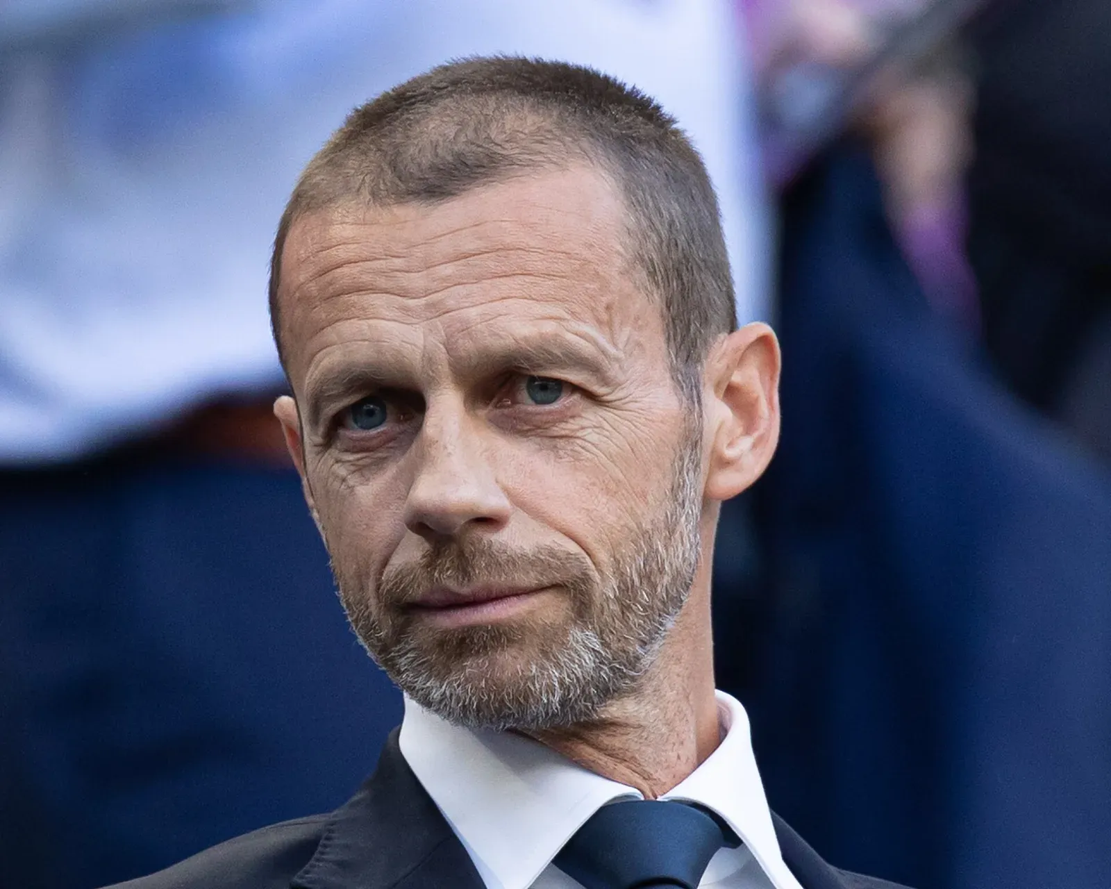 Aleksander Čeferin, de traje oscuro, camisa blanca y corbata azul, mira al frente con gesto serio ante un fondo desenfocado de público.