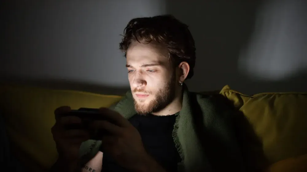 Un hombre barbudo jugando videojuegos en el interior, enfocado en una consola de mano.