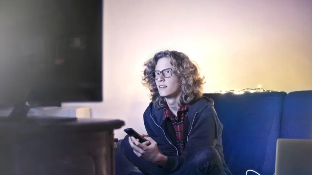 Concentrated male with long hair sitting on comfortable sofa at home and messaging on social media via cellphone while watching movie on TV with opened mouth