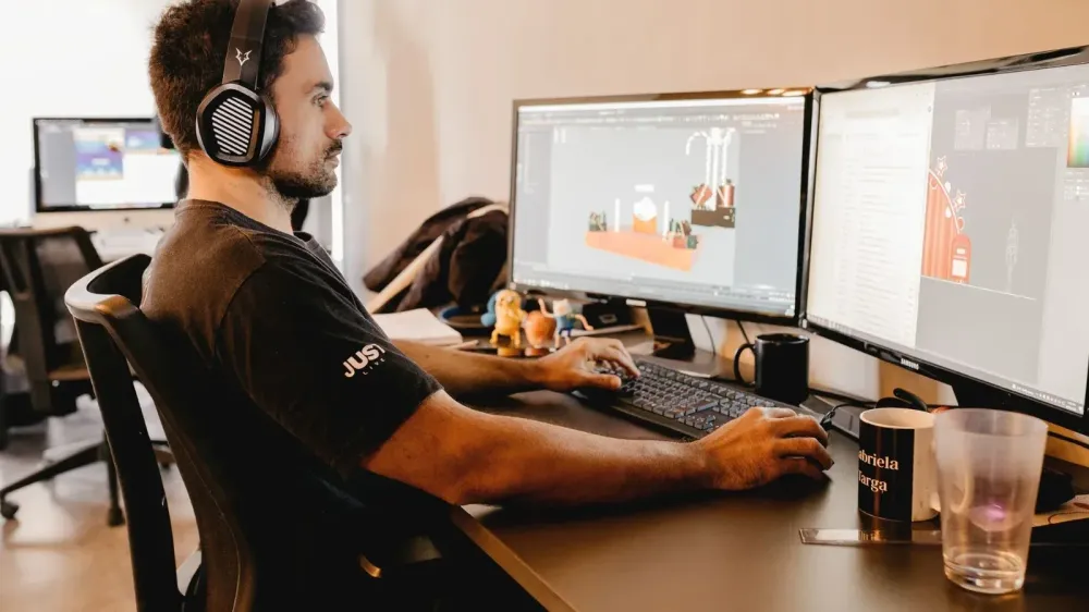 A man wearing headphones works on a computer with two monitors in a modern office setup.