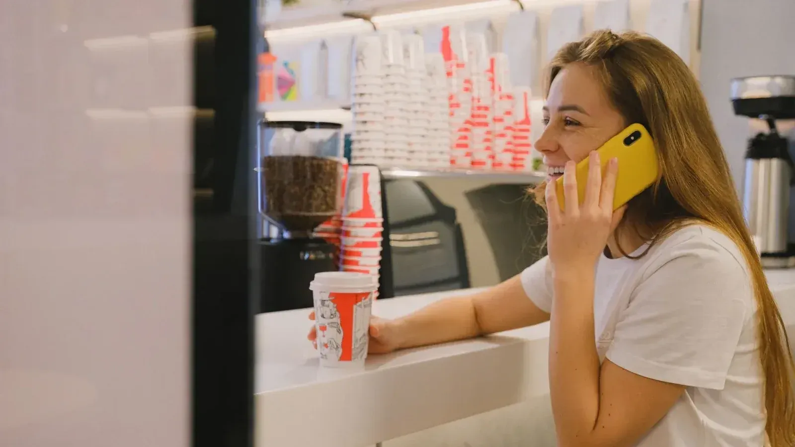 Smiling woman in coffee shop talking on smartphone, enjoying a hot drink.