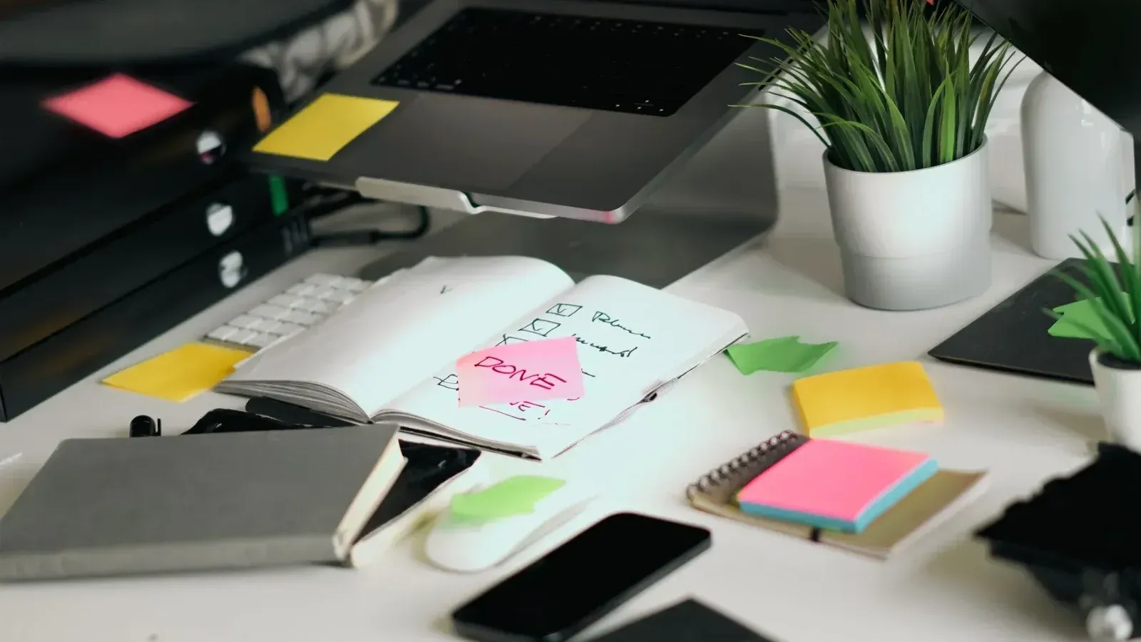 A cluttered office desk featuring a laptop, notebooks, and checklists amid colorful sticky notes.