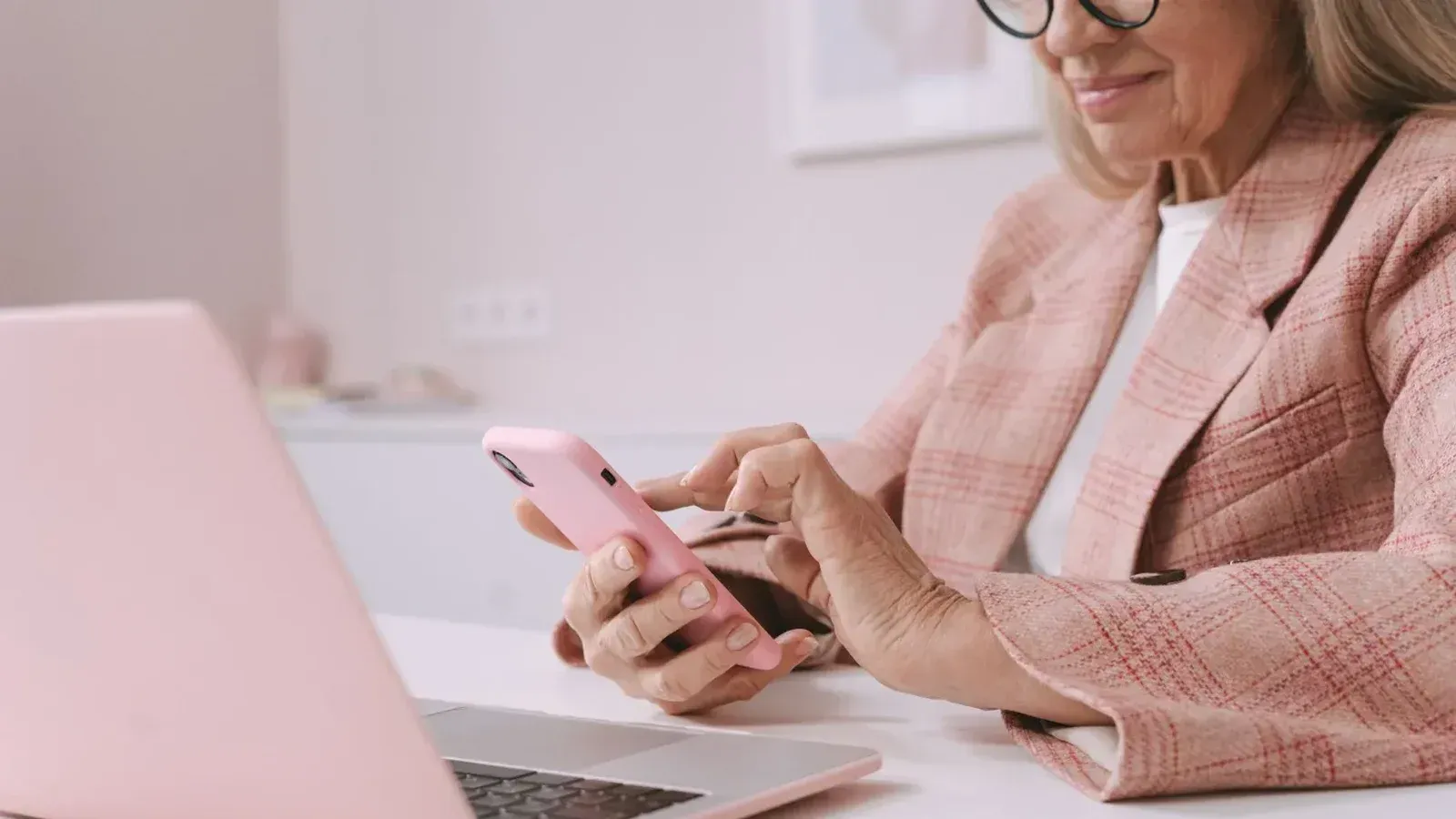 Anciana en traje de cuadros rosa con un smartphone y una computadora portátil en un escritorio.