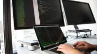 A developer writes code on a laptop in front of multiple monitors in an office setting.