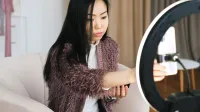 An Asian woman vlogs at home using a smartphone and ring light, perfect setup for influencers.