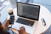 A programmer manages code on a laptop while multitasking with a smartphone, illustrating modern tech workspace.
